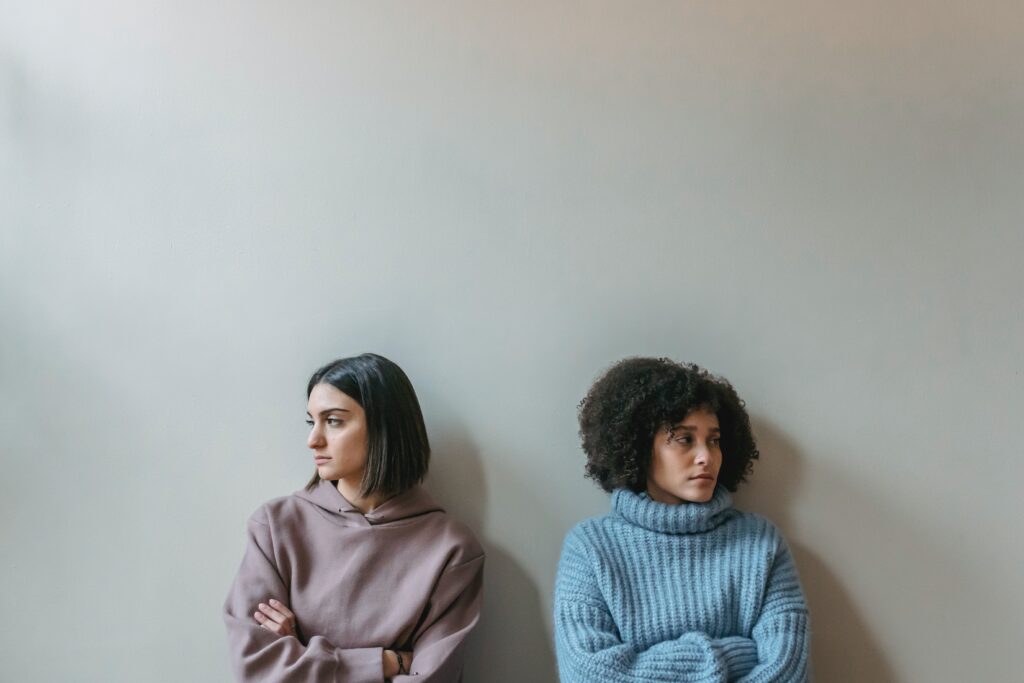 two women sit apart against a wall with arms crossed and heads turned in opposite directions